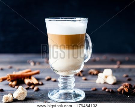 Hot latte macchiato coffee with tasty foam in tall clear glass on dark wooden table serving with cinnamon, cane sugar and roasted coffee beans. Breakfast time. Low key photo.