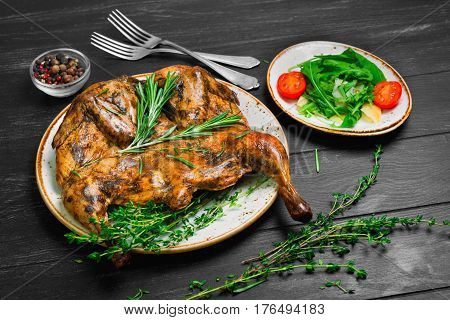 Georgian cuisine. Grilled fried roast Chicken Tapaka (Tabaka tobacco) in plate on black wooden background. Ingredients for Chicken arugula cheese thyme rosemary pepper cherry tomatoes.