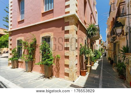 Rethymnon Island Crete Greece - June 23 2016: The narrow street of the old town's part of city Rethymnon and red colored house with plants and flowers in the big pots in hot sunny summer day.