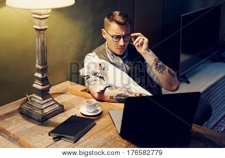 Handsome tattooed man in eyeglasses working at home on laptop while sitting at the wooden table with cute dog on his hands.Adult guy using modern computer for research new business ideas