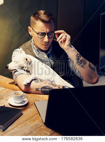 Handsome tattooed man in eyeglasses working at home on laptop while sitting at the wooden table with cute dog on his hands.Adult guy using modern computer for research new business ideas.Vertical