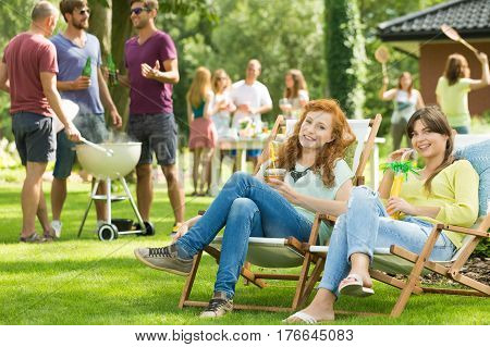 Two girls sitting on deckchairs with drinks men grilling