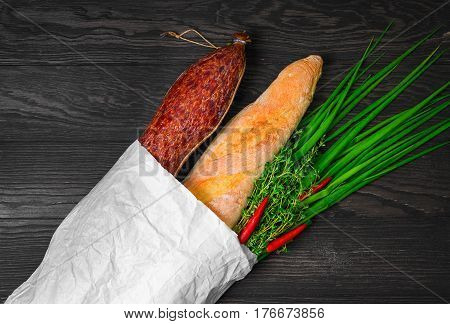 A grocery shopping set in a paper bag at the grocery store for sandwiches. Smoked sausage bread baguette green onion thyme red pepper. Dark brown wooden background.
