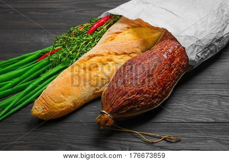 A grocery shopping set in a paper bag at the grocery store for sandwiches. Smoked sausage bread baguette green onion thyme red pepper. Dark brown wooden background.