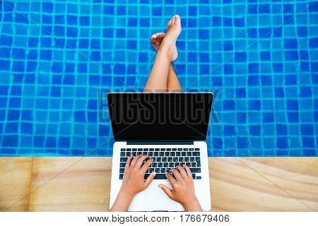 Woman working on her laptop computer sitting at poolside. Top view of female hand on the keyboard feet in the pool.