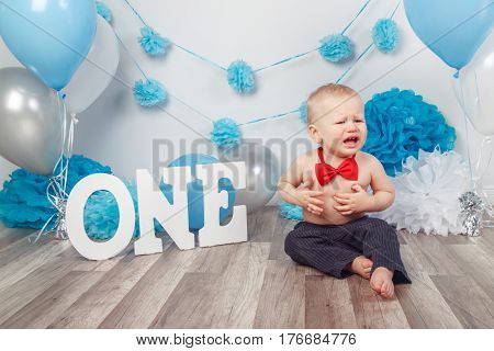Portrait of adorable Caucasian baby boy with blue eyes in dark pants and red bow tie celebrating his first birthday with letters one and balloons sitting on wooden floor in studio