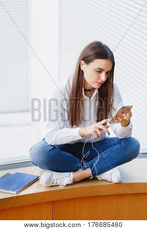 Portrait of young beautiful stylish young Caucasian girl woman in white shirt and blue jeans sitting on window sill in college university watching listening to lecture or music on her phone