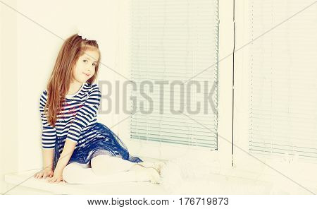 Little girl in blue striped dress and a white bow on her head.She sits at the window turned away from the camera.Creative toning of a photograph.