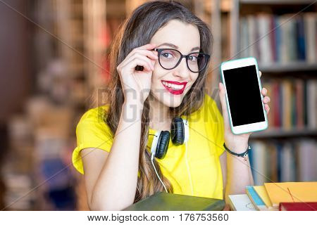 Young smiling student showing smart phone with empty screen standing at the library. Mobile application concept