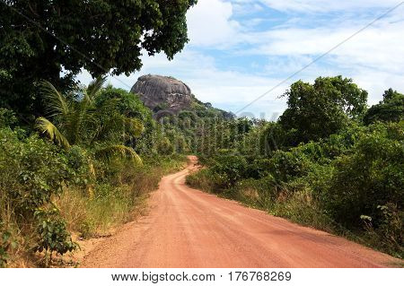 Dusty road going through green colored bushes leading to a big black rock at midday and nobody around on the island Belitung Indonesia.