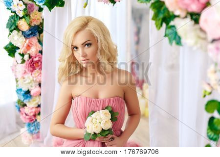 Young blond woman wearing pink gorgeous dress near the flowers. Horizontal indoors shot.