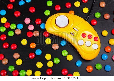 Video game console GamePad on a white wooden table. Yellow retro gamepad on a background of colored chocolate dragees. Copy space.