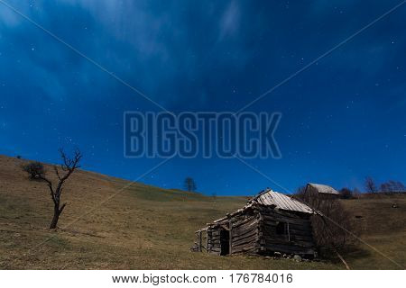 Shattered wooden cottage on top of the hill under the blue sky with lots of stars. Night landscape in Carpathians Romania