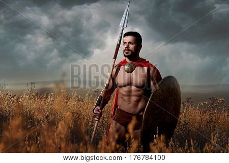 Athletic and handsome warrior with beard in red cloak with sword and shield going in attack. Soldier with bare torso like spartan or antique roman looking away. Dark sky over field.