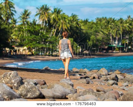Young woman walking on tropical beach. Pretty girl walks on beach. Romantic seashore blue sea and coco palm trees. Summer vacation casual photo. Relaxed day by the sea. Holiday leisure concept image