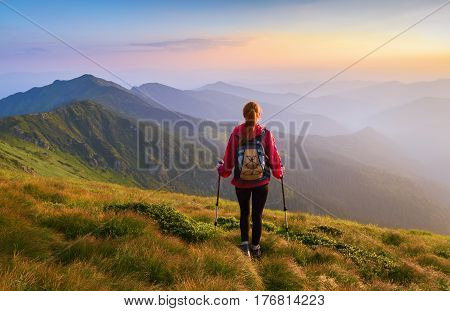 Redheaded girl athlete with a backpack and sticks stands on the green hillocks and looks at high mountain landscapes and fascinating sunset.