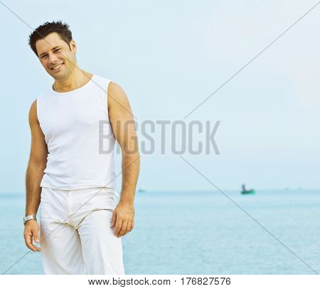 young brunette man on beach at sea smiling, gomosexual in white dress big muscles