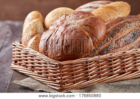 Wicker basket with bread. Bread and buns inside basket. Fresh bakery products on table. Tastes best when warm. On the wooden table with flour. Brown background. Bread on the cloth linen.