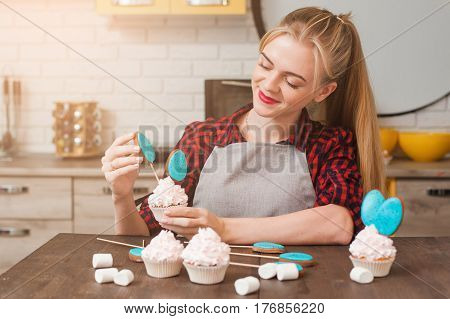 Girl decorating cup cakes with white cream and blue cake-pops at kitchen wooden table. Culinary masterclass. Easter gift, small business, delivery of sweets, craftmanship concept