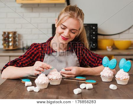 Girl decorating cup cakes with white cream and blue cake-pops at kitchen wooden table. Culinary masterclass. Easter gift, small business, delivery of sweets, craftmanship concept