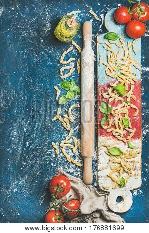 Ingredients for cooking Italian dinner. Fresh pasta casarecce, tomatoes, basil leaves, bottle of olive oil on colorful wooden board over dark blue background. Top view, copy space. Food frame concept