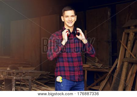Smiling Workman Dressed In The Checkered Shirt Wearing Protective Headphones At The Sawmill. Timbers