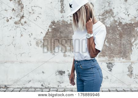 Hipster girl wearing blank white t-shirt, jeans and baseball cap posing against rough street wall, minimalist urban clothing style