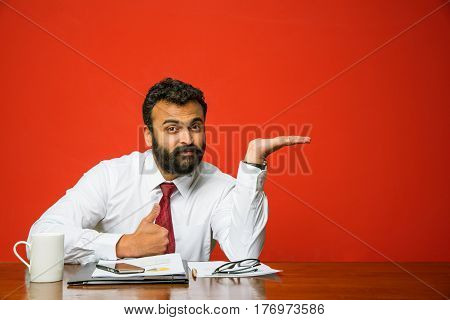 Bearded indian businessman sitting at table and presenting something with left hand, pointing towards blank wall with copyspace, asian business person presenting something