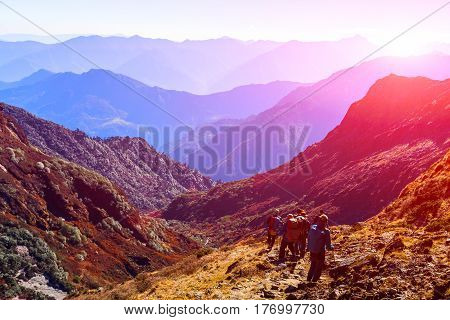 Group of People walking on Trek carrying Backpacks using trekking Sticks dressed in alpine Jackets and hiking Pants. Layered Mountains View with shining Sun beside