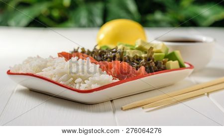 Bowl With Rice, Avocado, Salmon And Kelp On A White Wooden Table Against A Green Tree. Dier Food.