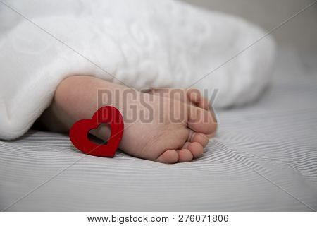 The Baby Sleeps Under A Blanket, Feet Out With A Red Heart For The Holiday