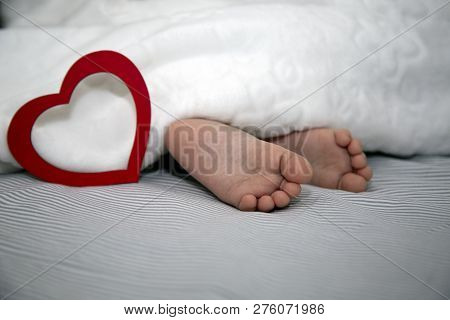 The Baby Sleeps Under A Blanket, Feet Out With A Big Red Heart For The Holiday