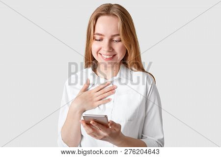 Studio Shot Of Pleased Young Woman Recives Message With Impressive Words, Keeps Hand On Chest, Smile