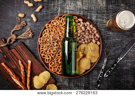 A Bottle Of Beer On A Plate With Salted Ookies Pretzels, Pistachio Nuts And Chips On A Black Scratch