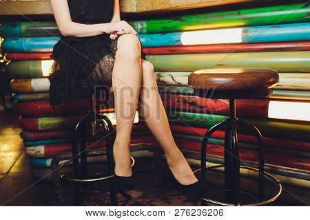 A Close Up Image Of The Legs Of A Young Woman In A Dress Sitting Ona Barchair In Black Heels.