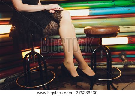 A Close Up Image Of The Legs Of A Young Woman In A Dress Sitting Ona Barchair In Black Heels.