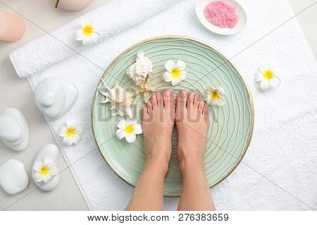 Woman Soaking Her Feet In Plate With Water, Flowers And Seashells On White Towel, Top View. Spa Trea