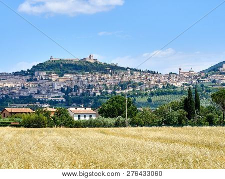 View Of The City Of Assisi, The Slope Of The Mountain Monte Subasio, Summer Clouds, Blue Sky