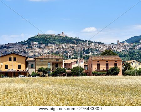 View Of The City Of Assisi, The Slope Of The Mountain Monte Subasio, Summer Clouds, Blue Sky