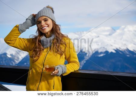 Woman drinking warm tea in the rustick wooden terrace on mountain, alpine view, snow on hills. 