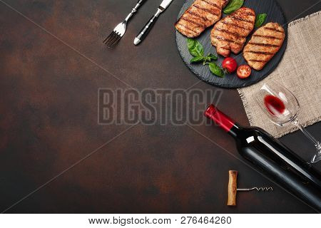 Grilled Pork Steaks On Stone With Bottle Of Wine, Wine Glass, Knife And Fork On Rusty Background