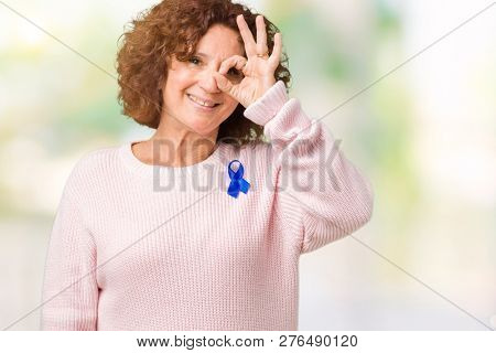 Middle ager senior woman wearing changeable blue color ribbon awareness over isolated background doing ok gesture with hand smiling, eye looking through fingers with happy face.