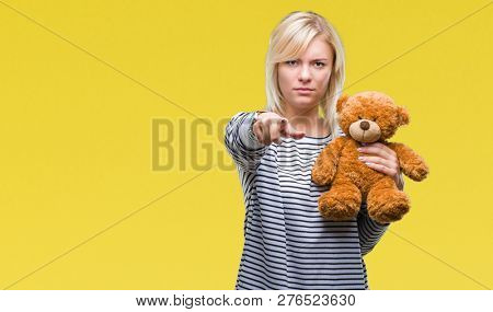 Young beautiful blonde woman holding teddy bear plush over isolated background pointing with finger to the camera and to you, hand sign, positive and confident gesture from the front