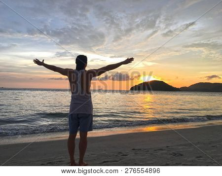 Man meditating whilst raising his arms an looking out to sea at the beautiful sunset on Pantai Cenang on Langkawi Island in Malaysia