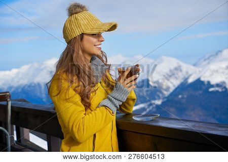 Woman drinking warm tea in the rustick wooden terrace on mountain, alpine view, snow on hills. 