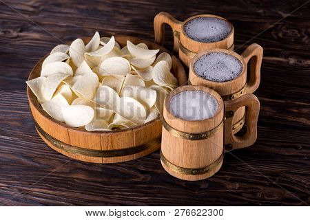 Three Oak Mugs With Beer Inside. They Stand Near An Oak Tray In Which Are Potato Chips. On Dark Wood