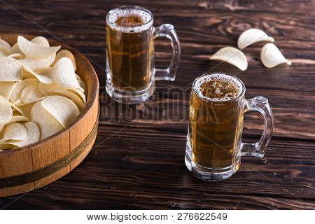 Two Glass Mugs Of Beer Stand On A Wooden Table Near An Oak Tray With Potato Chips Inside