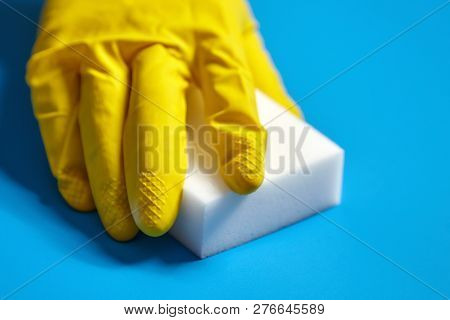 Human Hand In Yellow Glove Holds A White Melamine Sponge On Blue Background