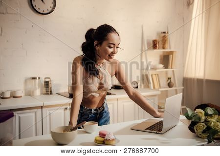 Beautiful Smiling Young Woman In Lace Bralette And Jeans Using Laptop During Breakfast At Home