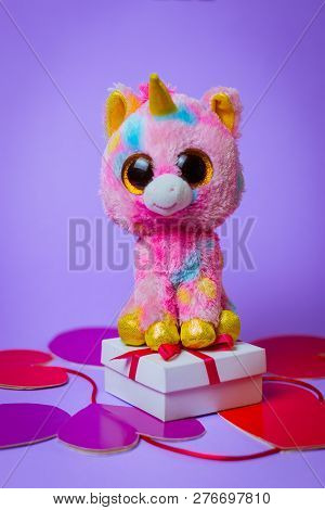 Toy Pink Unicorn Sits On A White Gift Box With A Red Ribbon On Paper Hearts On A Purple Background